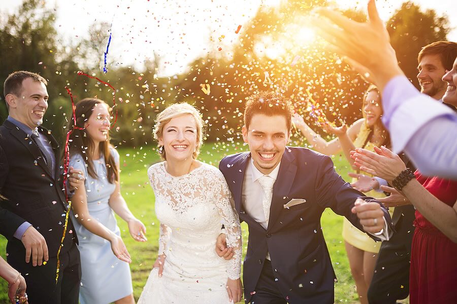 Full length portrait of newlywed couple and their friends at the wedding party showered with confetti in green sunny park.