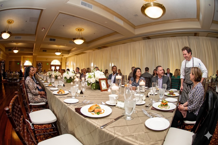 People sitting at dinner tables at a conference while speaking to the chef.